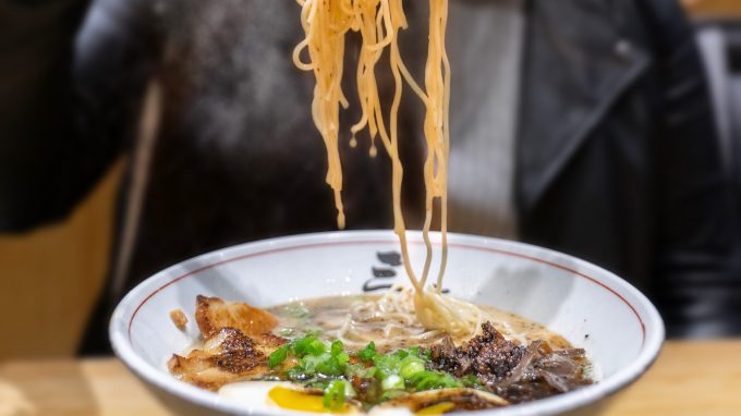 Noodles being picked up over steaming bowl of orange ramen broth.